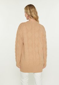 Beige gestrickte Strickjacke mit einem Zopfmuster, oversized Schnitt, gerippte Bündchen und Saum. Glatte Textur, mittellanges Design.