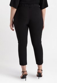 Mustat slim-fit housut, joissa on joustava vyötärö, lyhyempi pituus ja hienovarainen kiilto, yhdistettynä ruskeisiin leopardikuosisilla korkokengillä.