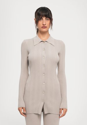 Frau mit dunklem Haar in einem gerippten, langärmeligen, knopfgebundenen beigen Cardigan und passenden Hosen, die vor einem schlichten weißen Hintergrund steht.