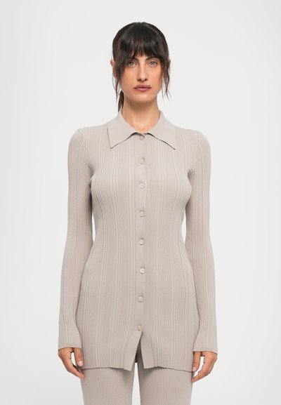 Femme aux cheveux foncés portant un cardigan beige à rayures, à manches longues et boutonné, ainsi que des pantalons assortis, se tenant devant un fond blanc uni.