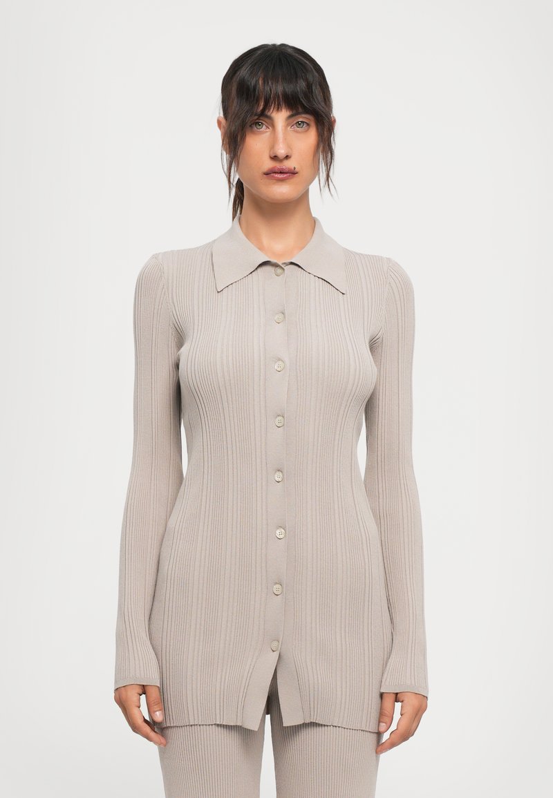 Woman with dark hair in a ribbed, long-sleeve, buttoned beige cardigan and matching pants, standing against a plain white background.
