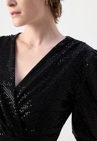 Robe noire avec un décolleté en V profond, tissu texturé avec des paillettes et des manches bouffantes froncées. Surface lisse et brillante avec des accents scintillants.