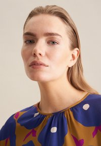 Blusa blu e oro con motivo floreale e dettagli bianchi. Presenta un colletto arricciato e una texture di tessuto liscia.