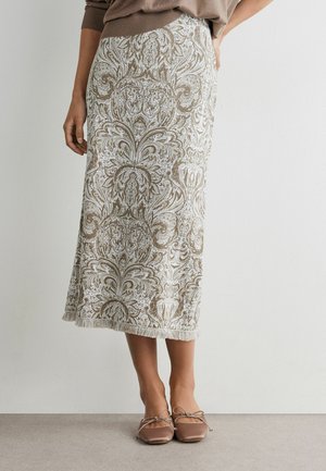 Frau trägt einen knielangen Rock mit filigranen weißen und braunen Blumenmustern, kombiniert mit braunen Ballerinas und einem braunen Oberteil.