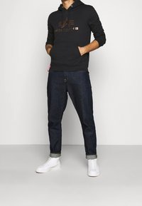 Sudadera negra con capucha con un logo metálico, bolsillos delanteros, combinada con vaqueros oscuros remangados y zapatillas blancas. Diseño sencillo y casual.