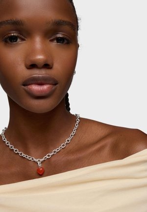 Primer plano de una persona que lleva un collar de cadena plateada con un pequeño colgante de corazón rojo y una blusa beige de hombros descubiertos sobre un fondo liso.