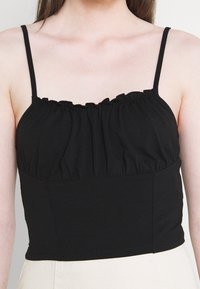 Černý bezramínkový top s volánkovým výstřihem, nařaseným materiálem na prsou a přiléhavou spodní částí. Hladká textura, minimalistický design.