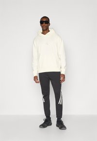 Sweatshirt com capuz em creme claro com um padrão texturizado e logótipo branco. Calças de fato cinza-escuro com detalhes laterais em branco. Sneakers pretos.