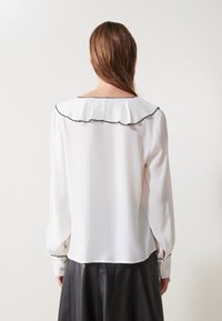 Vit blus av lätt tyg med en ryschkrage kantad i svart, långa ärmar och en lös passform, kombinerad med en svart läderkjol.