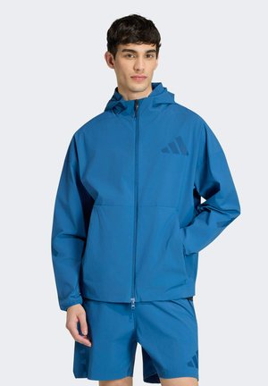 Joven con chaqueta azul con capucha y cremallera y pantalones cortos a juego con un logotipo sutil, de pie frente a un fondo claro y liso.