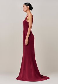 Burgundska oprijeta obleka z okroglim izrezom, dolžine do tal in gumbi ob strani. Izdelana je iz gladkega, elastičnega materiala.