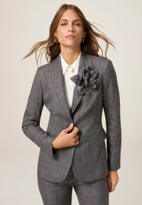 Vrouw in een grijs ruitpak met rode lijnen, witte blouse en grote stoffen bloemenpin op de revers van het blazer, poserend tegen een effen achtergrond.