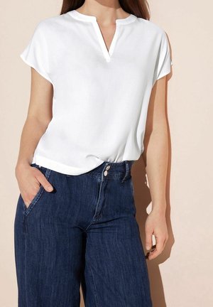 Frau trägt eine weiße kurzärmelige Bluse, die in hoch taillierte dunkelblaue Jeans aus Denim gesteckt ist, mit der Hand in der Tasche.