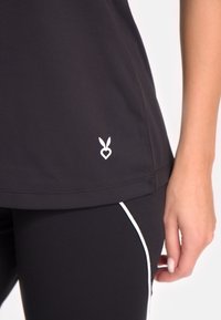 Melns sporta tops ar gludu tekstūru, ar baltu sirdi un trušu ausīm logotipu apakšmalā. Saskaņots ar melnām legingām, ko akcentē baltas svītras.