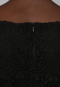Czarna koronkowa sukienka z tyłu, z misternymi wzorami kwiatowymi, gładkim zamkiem błyskawicznym i falistym dekoltem o teksturowanym wykończeniu.