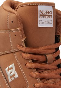 Zapatilla alta marrón con parte superior de cuero suave, logotipo blanco en contraste y cordones a juego. Presenta un cuello acolchado y acentos cosidos.