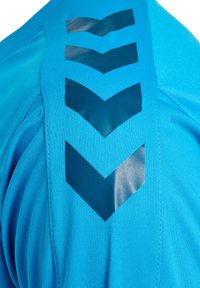 Zbliżenie na niebieską sportową koszulkę z czterema ciemnoniebieskimi skośnymi paskami skierowanymi w dół na ramionach.