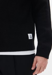 Suéter negro de punto acanalado con un patrón texturizado, con una etiqueta blanca en el dobladillo marcada como "M 73". Lleva puesto sobre una camisa blanca.