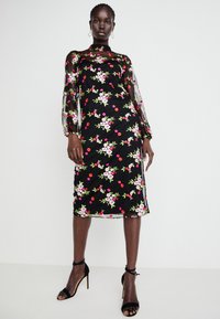 Vestido preto com bordado floral em rosa e vermelho. Mangas longas transparentes, corte justo, até ao joelho, combinado com sandálias de salto alto pretas.