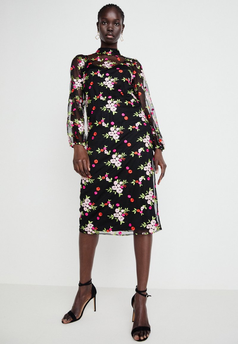 Crna haljina s cvjetnim vezom u ružičastoj i crvenoj boji. Prozirne duge rukave, uski kroj, do koljena, uparena s crnim sandalama na visoku petu.