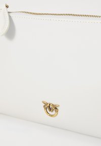 Witte leren handtas met een gouden hardwaredetail met een gevleugeld ontwerp. Inclusief een ritssluiting langs de bovenrand.