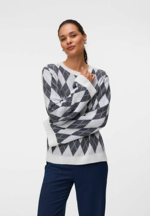 Kvinde iført en grå og hvid rombemønstret sweater og mørkeblå bukser, der poserer med armene krydset foran en ensfarvet hvid baggrund.