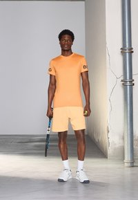 Πορτοκαλί αθλητικό μπλουζάκι με μαύρο λογότυπο "BG", συνδυασμένο με ανοιχτό πορτοκαλί σορτσάκι. Το μοντέλο κρατάει μια ρακέτα τένις και μια πράσινη μπάλα τένις.