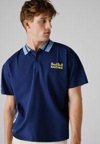Námořnická polo tričko se světle modrým límečkem. Na přední straně je žluté logo "Red Bull RACING". Krátké rukávy, hladká tkanina.