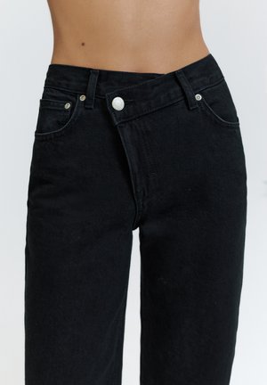 Zwarte denim jeans gedragen op de taille met een asymmetrische tailleband en knoopsluiting, waarbij de buik zichtbaar is tegen een effen achtergrond.