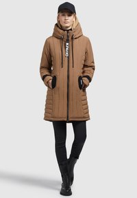 Vrouw in een bruin gewatteerd hooded jack met zwarte patches op de zakken, zwarte pet, zwarte broek en zwarte enkellaarzen, staand met handen in de zakken.