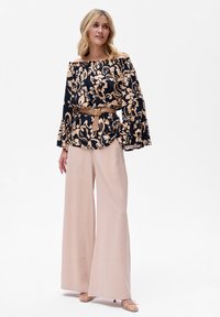 Kwiatowa bluzka bez ramiączek w odcieniach granatu i beżu, z rękawami dzwonami i paskiem w talii, połączona z szerokimi beżowymi spodniami i jasnymi butami.