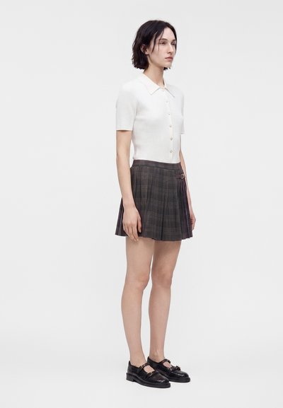 Femme aux cheveux courts et foncés, portant un haut blanc à manches courtes boutonné, une jupe plissée grise et des chaussures noires, debout devant un fond uni.