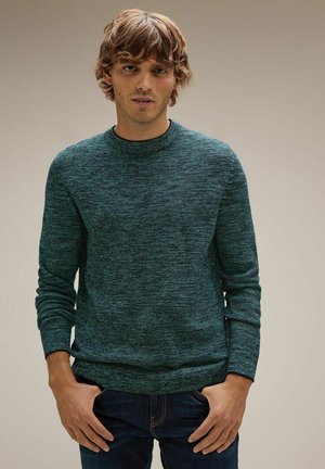 Grüner strukturierter Strickpullover mit Rundhalsausschnitt und langen Ärmeln, kombiniert mit blauen Jeans, vor einem neutralen Hintergrund präsentiert.