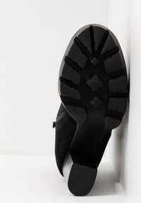 Bottines en daim noir avec une semelle en caoutchouc texturée et à motifs. Présente un talon épais et une fermeture éclair sur le côté. La semelle a des crampons profonds pour une bonne adhérence.