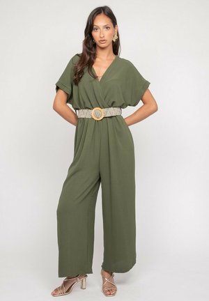 Femme portant une combinaison ample vert olive à manches courtes, une ceinture à motifs et des talons beiges à brides, debout les mains sur les hanches.