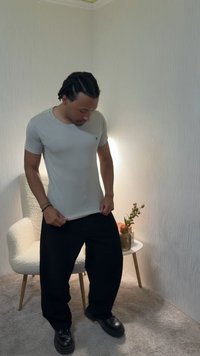 Άνδρας με γκρι κοντομάνικη μπλούζα και μαύρο παντελόνι στέκεται κοντά σε μία λευκή πολυθρόνα και ένα μικρό τραπέζι με φυτό σε ένα φωτεινό δωμάτιο με ελαφρώς υφασμένους τοίχους.