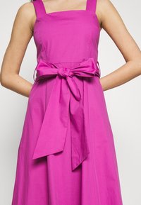 Fuksia puuvillamekko, jossa on neliönmuotoinen kaula-aukko, paksut olkaimet ja solmittava vyö, joka korostaa vyötäröä. Kellohame pehmeällä tekstuurilla.