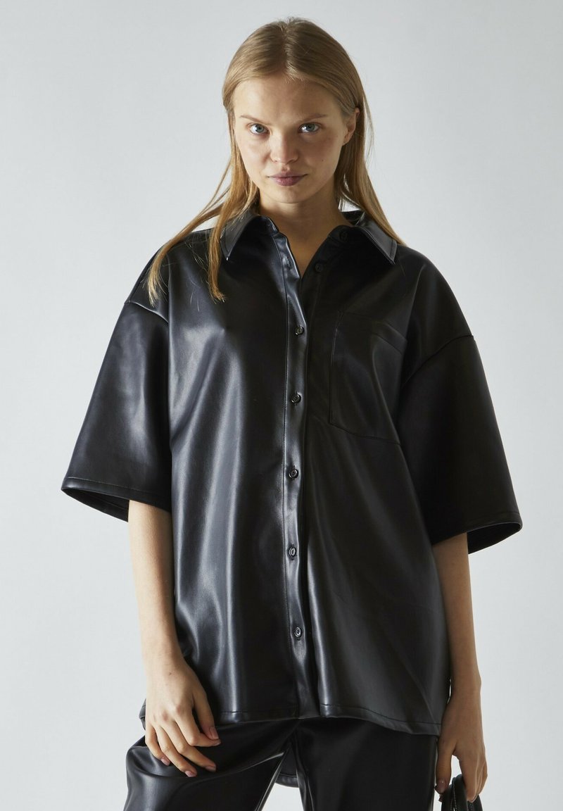 Camisa de cuero negro de botones con mangas cortas, que presenta un bolsillo en el pecho y una textura brillante, sobre un fondo neutro.