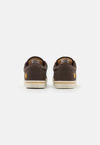 Sapatos de lona castanhos com um acabamento texturizado, solas brancas e acentos laranja no calcanhar, apresentando um colarinho almofadado e um detalhe de marca.