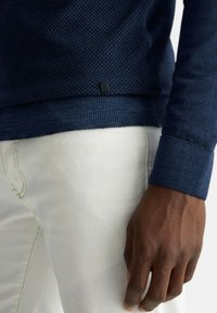 Maglione navy a trama con polsini a coste, etichetta del marchio visibile, indossato sopra pantaloni bianchi. Messa a fuoco sui dettagli del tessuto e sul contrasto di colore.