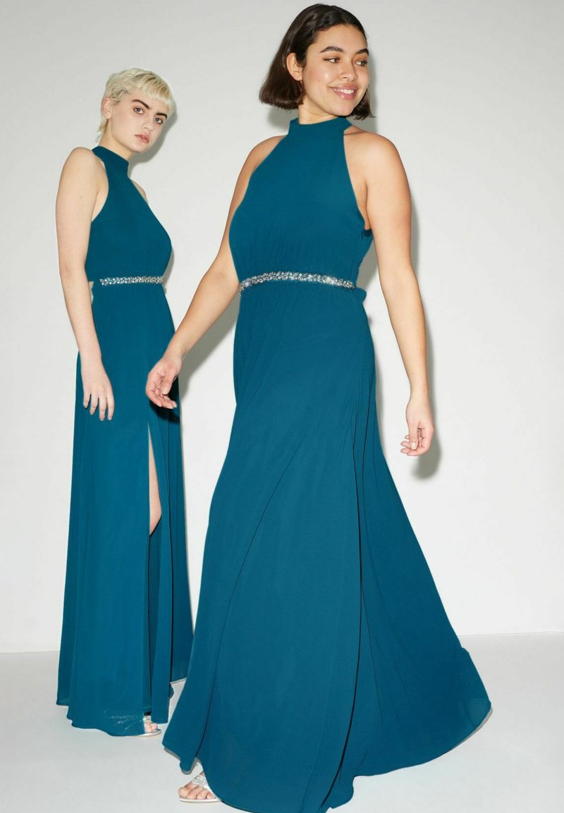 Due modelle indossano lunghi abiti color teal scuro con colli alti e gonne fluide. Ogni abito presenta una cintura di paillettes in vita e uno spacco laterale.