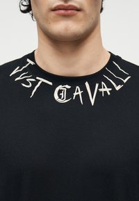 Κοντινό πλάνο ενός ατόμου που φοράει μαύρο πουκάμισο με λευκή στυλιζαρισμένη επιγραφή "Just Cavalli" γύρω από το γιακά.