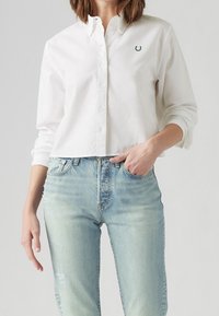 Chemise blanche boutonnée avec un col pointu, ornée d'un logo vert brodé, associée à un jean bleu clair déchiré.