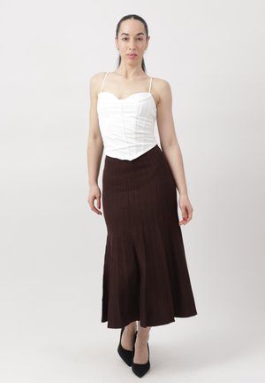 Kobieta stojąca na białym tle, ubrana w biały bezrękawnik typu gorset, ciemnobrązową plisowaną długą spódnicę i czarne szpilki.