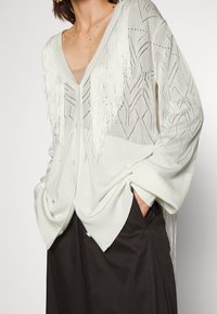 Femme portant un cardigan en maille blanche avec des détails à franges et des motifs géométriques, associé à un pantalon noir à jambes larges et les mains dans les poches.