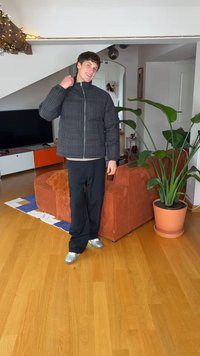 Μαύρο καπιτονέ μπουφάν με ψηλό γιακά, ασημένιο φερμουάρ και καρό σχέδιο. Μαύρο παντελόνι και μεταλλικά ασημί παπούτσια συμπληρώνουν την εμφάνιση. Εσωτερικός χώρος.