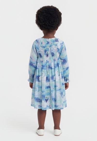 Copil cu păr creț purtând o rochie albastră cu mâneci lungi până la genunchi, cu modele de lebede, cu fața spre spate, stând pe un fundal alb.