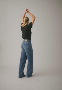 Kõrge vöökohaga sinised teksapüksid laia säärtega, tagumisel vöökohal tan-nahaline nahast detail ja kaks tagataskut. Seljas on must T-särk.