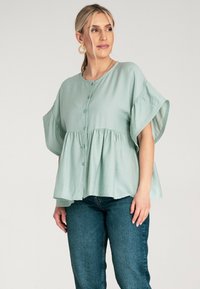Jasnozielona bluzka z krótkim rękawem, z zapięciem na guziki z przodu i marszczonym pasem. Miękka, teksturowana tkanina tworzy luźny fason. Zestawiona z ciemnymi dżinsami.