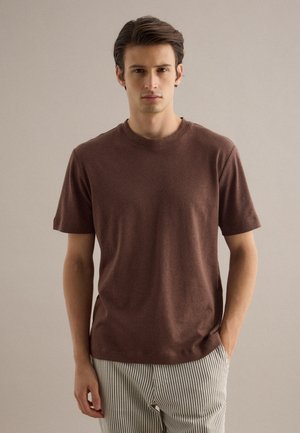 Giovane uomo con una camicia marrone a maniche corte e pantaloni a righe, in piedi con una mano in tasca contro uno sfondo semplice.